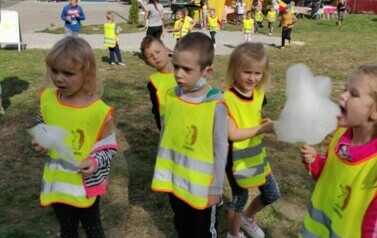 Dzieci stoją. Jedzą watę cukrową. Mają na sobie kamizelk odblaskowe.