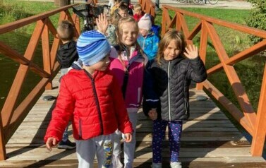 Dzieci idą w parach po drewnianym mostku. Machają do zdjcia. Uśmiechają się.