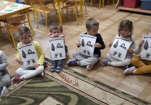 Dzieci siedzą na dywanie. Pokazują swoje prace plastyczne.