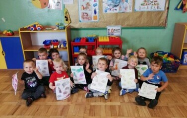 Dzieci siedzą na podłodze. W rękach trzymają prace plastyczne. Pokazują. Machają rękami do zdjęcia.