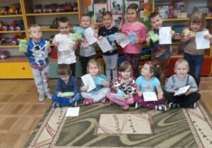 Zdjęcie grupowe. Dzieci trzymają w rękach prace plastyczne. Za nimi jest tablica z uszytą szczoteczką, pastą do zęb&oacute;w i mydłem