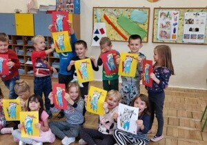Zdjęcie grupowe. Dzieci trzymają w rękach prace plastyczne. Za nimi jest tablica z uszytą szczoteczką, pastą do zęb&oacute;w i mydłem