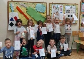 Zdjęcie grupowe. Dzieci trzymają w rękach prace plastyczne. Ubrane są na galowo z okazji Dnia Czyścioszka. Za nimi jest tablica z uszytą szczoteczką, pastą do zęb&oacute;w i mydłem