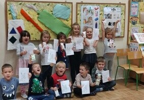 Zdjęcie grupowe. Dzieci trzymają w rękach prace plastyczne. Ubrane są na galowo z okazji Dnia Czyścioszka. Za nimi jest tablica z uszytą szczoteczką, pastą do zęb&oacute;w i mydłem.