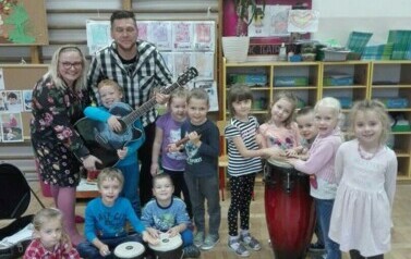 Zdjęcie grupowe. Dzieci uśmiechają się. Stoi z nimi nauczycielka i nauczyciel. Nauczyciel trzyma gitarę. Przed dziećmi stoją dwa bębenki