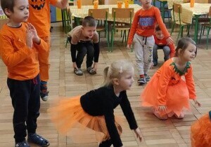Dzieci ubrane są na pomarańczowo. Dziewczynka kuca. Dzieci stoją w rozsypce.