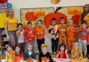 Zdjęcie grupowe. Dzieci ubrane są na pomarańczowo. Za nimi na tablicy wisi dynia. Z dziećmi stoją nauczycielki.