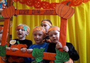 Dzieci na głowach mają pomarańczowe opaski. Zaglądają przez ramkę z napisem Dzień Dyni.