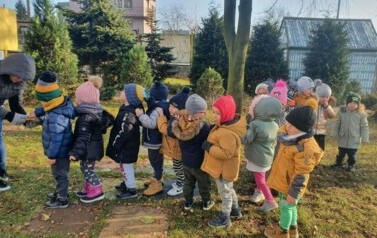 Dzieci ubrane są w kurtki i czapki. Stoją w rzędzie. Nauczycielka nasypuje dziecku na rękę karmę dla ptak&oacute;w.