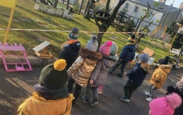 Dzieci ubrane są w kurtki i czapki. Są przed przedszkolem. Idą w kierunku karmnik&oacute;w. Karmniki stoją na krawężniku.