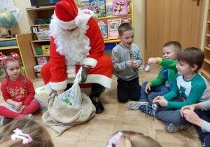 Dzieci siedzą na dywanie. Wśr&oacute;d nich na krzesełku siedzi święty Mikołaj. Wyjmuje coś z worka. Dzieci się cieszą.