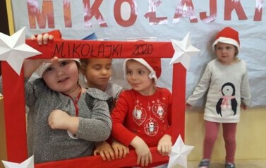 Tr&oacute;jka dzieci zagląda do czerwonej ramki. Na ramce jest napisane Mikołajki 2020. Dzieci mają czapki mikołajkowe na głowach.