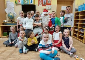 Zdjęcie grupowe. Dzieci stoją i siedzą w sali przedszkolnej na dywanie w grupie. Pośr&oacute;d nich siedzi święty Mikołaj.
