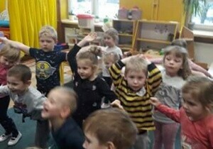 Dzieci stoją w rozsypce na dywanie w sali przedszkolnej. Pokazują gesty. 