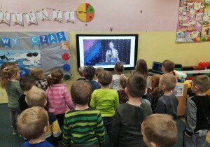 Dzieci stoją w rozsypce na dywanie w sali przedszkolnej. Patrzą na ekran. 