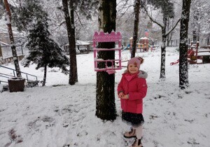 Dziewczunka stoi przy karmniku w ogrodzie przedszkolnym. Uśmiecha się do zdjęcia.Na podw&oacute;rku jest śnieg. Ubrana jest w kurtkę, czapkę, rękawiczki.