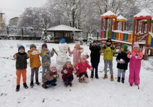 Zdjęcie grupowe w ogrodzie przedszkolnym. Na podw&oacute;rku jest śnieg. Dzieci uśmiechaja się. Pośr&oacute;d nich stoi bałwan ulepiony ze śniegu. Dzieci ubrane są w kurtki, czpki, szaliki i rękawiczki.
