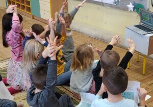 Dzieci siedzą na dywanie w sali przedszkolnej. Podnoszą ręce do g&oacute;ry.