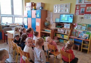 Dzieci siedzą na krzesełkach przed telewizorem. Oglądają prezentację multimedialną o ekologii.