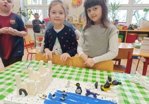 Dwie dziewczynki stoją przy stoliku. Uśmiechają się do zdjęcia. Przed nimi stoi makieta Antarktydy.