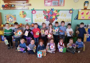 	 Zdjęcie grupowe dzieci. Dzieci są odświętnie ubrane. Za nimi wisi tablica przygotowana na Dzień Babci i Dziadka. Uśmiechają się do zdjęcia. W rękach trzymają laurki.