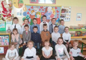 	 Zdjęcie grupowe dzieci. Dzieci są odświętnie ubrane. Za nimi wisi tablica przygotowana na Dzień Babci i Dziadka. Uśmiechają się do zdjęcia.