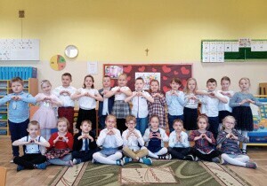 	 Zdjęcie grupowe dzieci. Dzieci są odświętnie ubrane. Za nimi wisi tablica przygotowana na Dzień Babci i Dziadka. Uśmiechają się do zdjęcia. W rękach trzymają laurki.