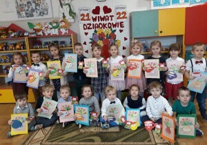 Zdjęcie grupowe dzieci. Dzieci są odświętnie ubrane. Za nimi wisi tablica przygotowana na Dzień Babci i Dziadka. Uśmiechają się do zdjęcia. W rękach trzymają laurki.