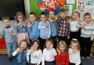 Zdjęcie grupowe dzieci. Dzieci są odświętnie ubrane. Za nimi wisi tablica przygotowana na Dzień Babci i Dziadka. Uśmiechają się do zdjęcia. Machają.
