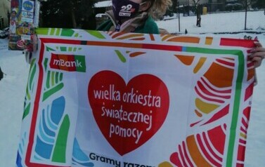 Dziewczyna trzyma plakat Wielkiej Orkiestry Świąteczenj Pomocy. Ma na twarzy maseczkę. 