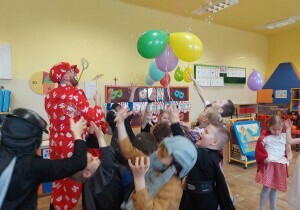 Dzieci są w sali przedszkolnej przebrane za r&oacute;żne postacie. Pośr&oacute;d nich stoi nauczyciel przebrany za klauna. W sali są kolorowe balony.