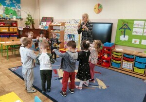 Nauczycielka stoi w klasie. Przed nią stoją dzieci.