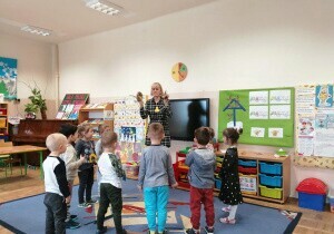 Nauczycielka stoi w klasie. Przed nią stoją dzieci.