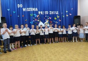Dzieci są odświętnie ubrane. Uśmiechają się do zdjęcia. Za nimi wisi dekoracja z napisem do widzenia przedszkole.