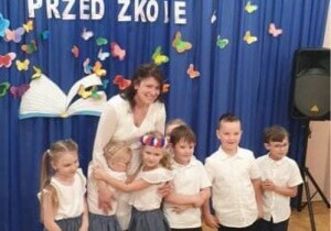Dzieci są odświętnie ubrane. Nauczyceielka stoi z dziećmi. Uśmiechają się do zdjęcia.