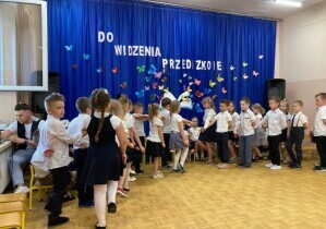 Dzieci tańczą poloneza. Są odświętnie ubrane. Za nimi wisi  dekoracja do widzenia przedszkola.