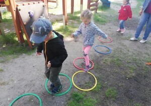 Dzieci są w ogrodzie przedszkolnym. Ubrane są w kurtki. Chłopiec z dziewczynką oprzechodzą z jednego koła do drugiego. Koła leżą na ziemi.