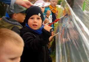 Dzieci malują farbami na streczu. Są ubrane w kurtki i czapki.