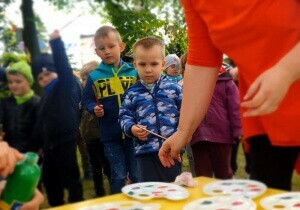 Dzieci są w ogrodzie przedszkolnym. Ubrane są w kurtki. Chłopiec patrzy na stolik. Na stoliku stoją podstawki z farbą.
