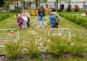 Dzieci stoją w dw&oacute;ch rzędach w ogrodzie przedszkolnym. Przed każdym rzędem stoi jedna bramka. Między rzędami stoi nauczycielka.