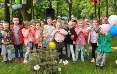 Dzieci stoją w grupie w ogrodzie przedszkolnym. Ubrane są w kurtki. Uśmiechają się do zdjęcia. Niekt&oacute;re dzieci trzymają baloniki.