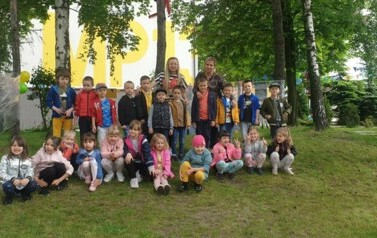 Dzieci stoją w grupie w ogrodzie przedszkolnym. Ubrane są w kurtki. Uśmiechają się do zdjęcia. Za nimi stoją nauczycielki.