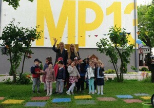 Dzieci stoją w grupie w ogrodzie przedszkolnym. Ubrane są w kurtki. Uśmiechają się do zdjęcia. Za nimi stoi nauczyciel.