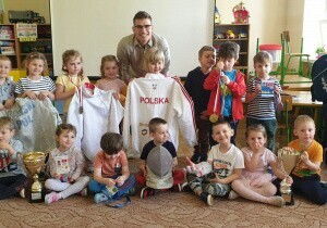 Zdjęcie grupowe. Dzieci siedzą lub stoją. Za nimi stoi pan. Dzieci trzymają w rękach bluze sportową z napisem Polska, puchary, maskę szermierską, medale.
