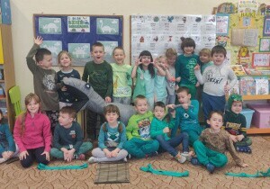 Zdjęcie grupowe. Dzieci ubrane są w zielone koszulki. Stoją lub siedzą na dywanie. Uśmiechają się do zdjęcia. 