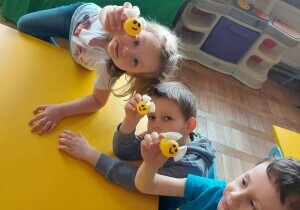 Dziewczynka i dw&oacute;ch chłopc&oacute;w siedzą przy stoliku. Pokazują plastikowe pszczoły wykonane z plastikowych jajek
