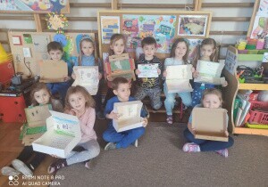 Zdkjęcie grupowe. Dzieci siedzą lub stoją w sali przedszkolnej. W rękach trzymają prace plastyczne.