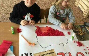 Chłopiec z dziewczynką siedzą przy stoliku. Wyklejają kontury Polski. Chłopiec uśmiecha się do zdjęcia.