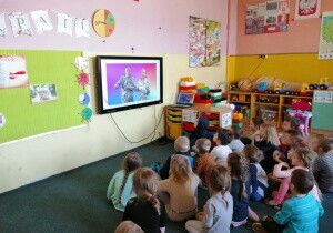 Dzieci siedzą na dywanie. Oglądają koncert na tablicy multimedialnej.