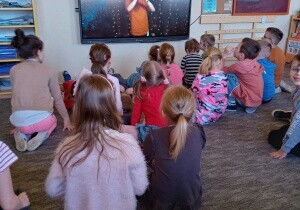 Dzieci siedzą na dywanie. Oglądają koncert na tablicy multimedialnej.
